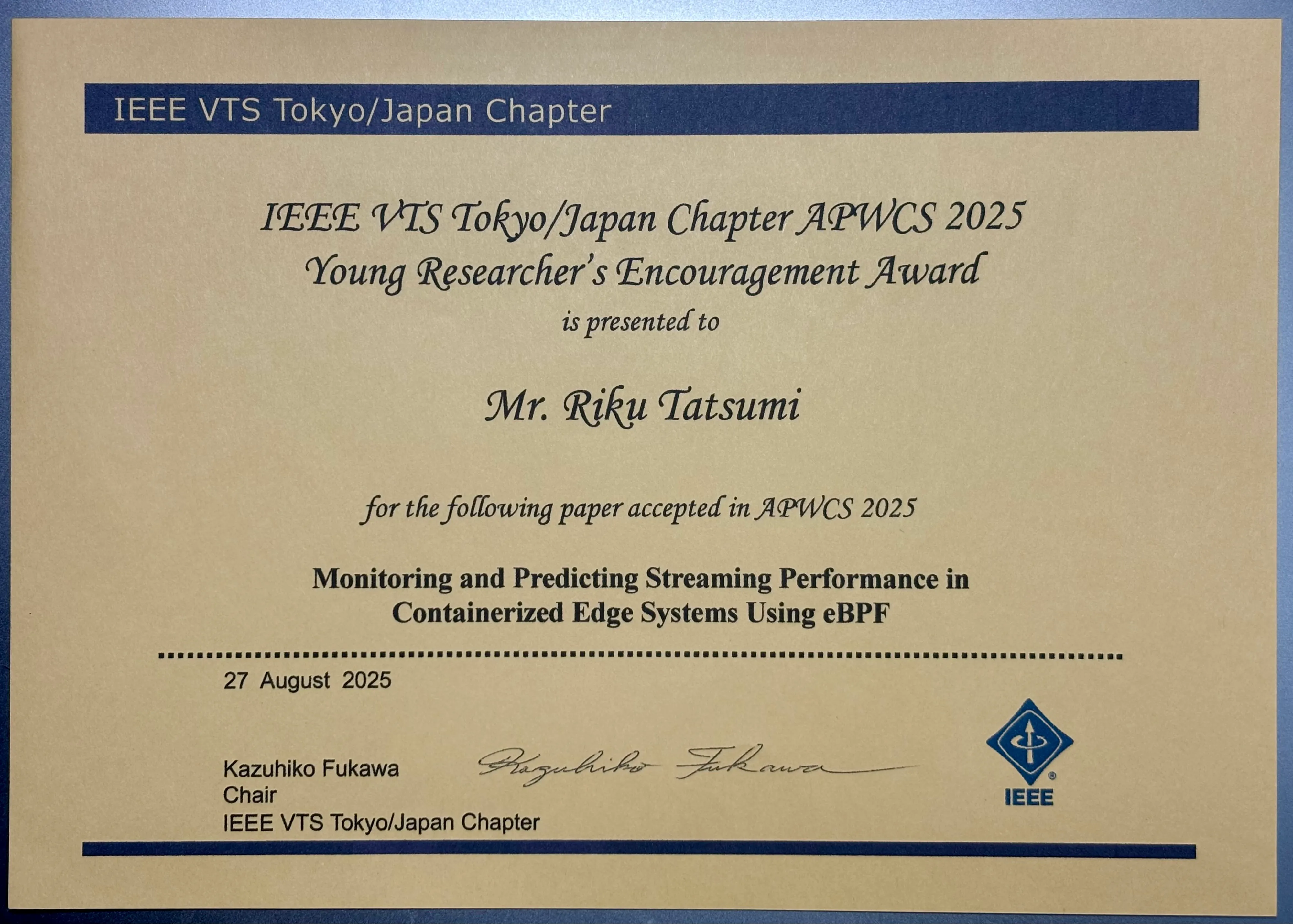 IEEE VTS Tokyo/Japan Chapter の「APWCS 2025 Young Researcher’s Encouragement Award（若手研究奨励賞）」の賞状。Mr. Riku Tatsumi に対し、APWCS 2025 採択論文「Monitoring and Predicting Streaming Performance in Containerized Edge Systems Using eBPF」に基づき授与された。日付は 2025年8月27日。署名は Kazuhiko Fukawa（Chair, IEEE VTS Tokyo/Japan Chapter）。右下に IEEE のロゴ。