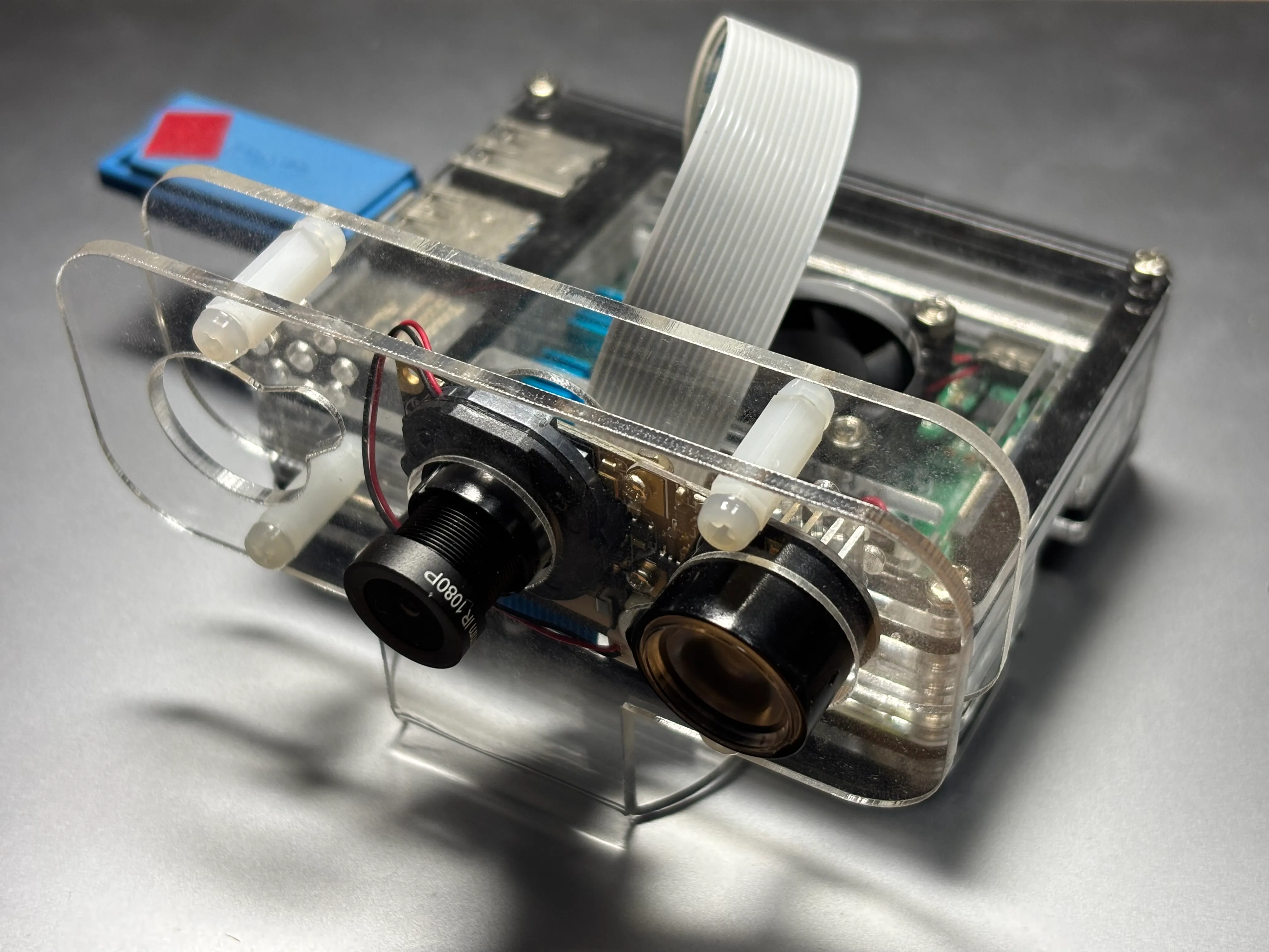 Raspberry Pi に2つのカメラモジュールを接続し、透明なアクリルケースに収めた自作装置。リボンケーブルで基板とカメラが接続され、内部には冷却ファンと電源モジュールが見える。